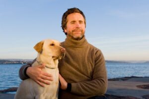 Blind mann ser mot kamera. Ved sin høyre side sitter en hund inntil ham. Hav i bakrunnen, lyset er aftensol.