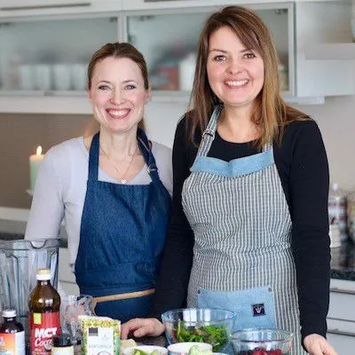 To kvinner med forkle, tatt i kjøkken, ingredienser til mat foran dem på bord.