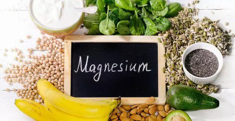 Sort tavle med hvit skrift, magnesium. Omsluttet av bananer, mandler, urter og korn, som er kilder til magnesium.