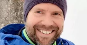 Mann som smiler, lue på hodet, ser i kamera, blå jakke.