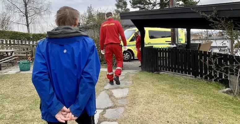 Eldre mann i blå jakke går med ryggen mot kamera. Forhan ham går to ambulansesjåfører kledd i rødt. I bakgrunnen er ambulansen.